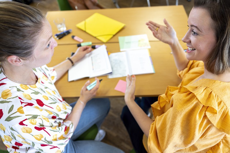 Two women in conversation with a booklet guide in the background and highlighters.