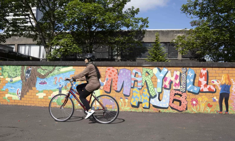 A woman on a bike cycling in front of a colourful wall, with the words 'Maryhill Hub' painted on.