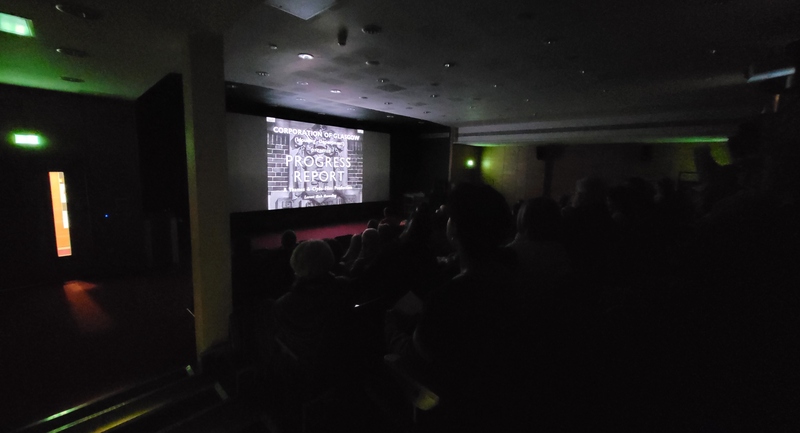 A dark cinema room with people watching the film on the screen. The title of the black and white film shown appears on the screen: 'Project Report'.