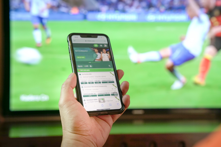 A tv with a football game on in the background, with a hand holding a phone with a betting app open in the foreground.